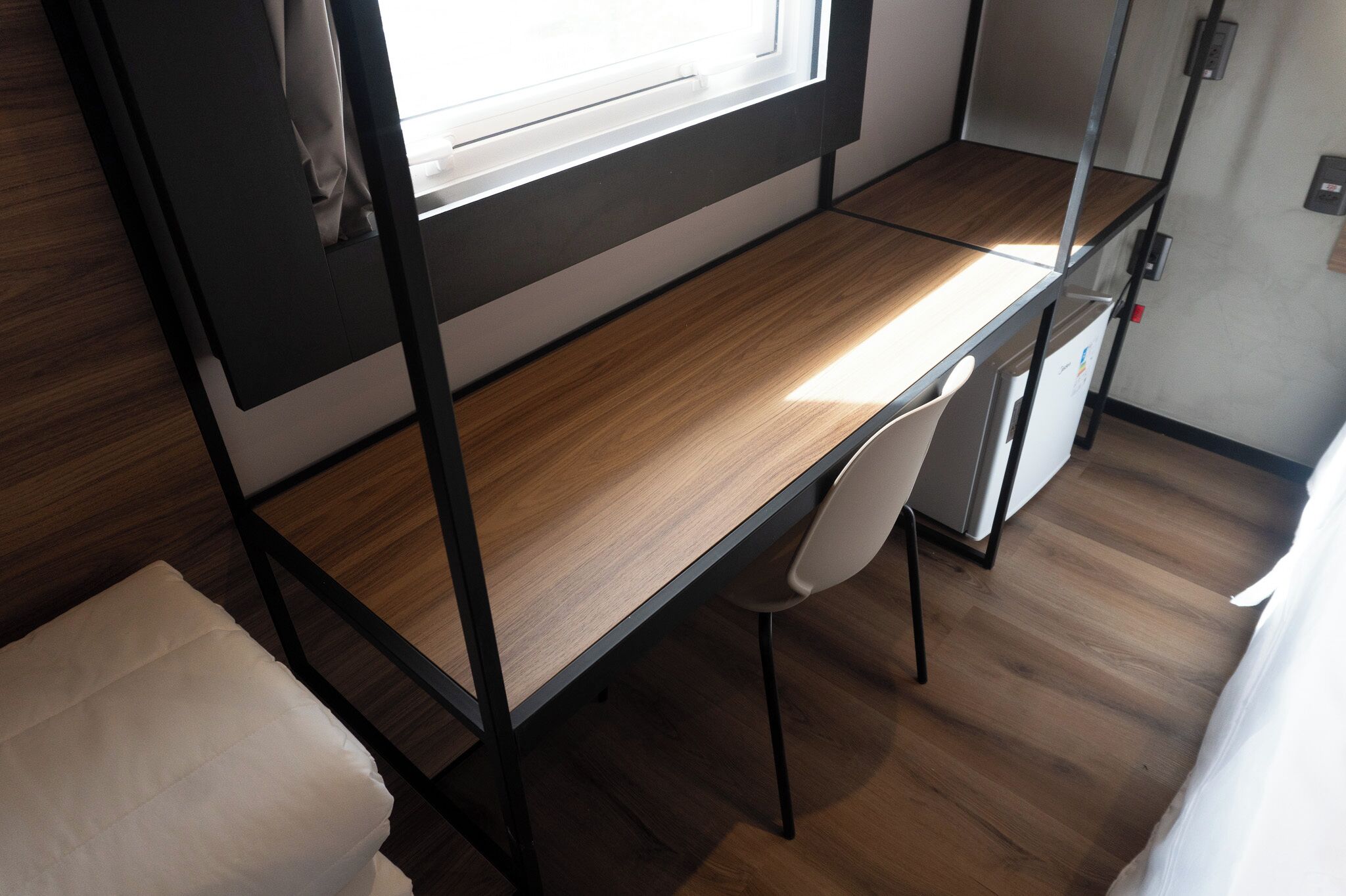 Een stapelbed met bureau en stoel, een klein raam en een wit gordijn.