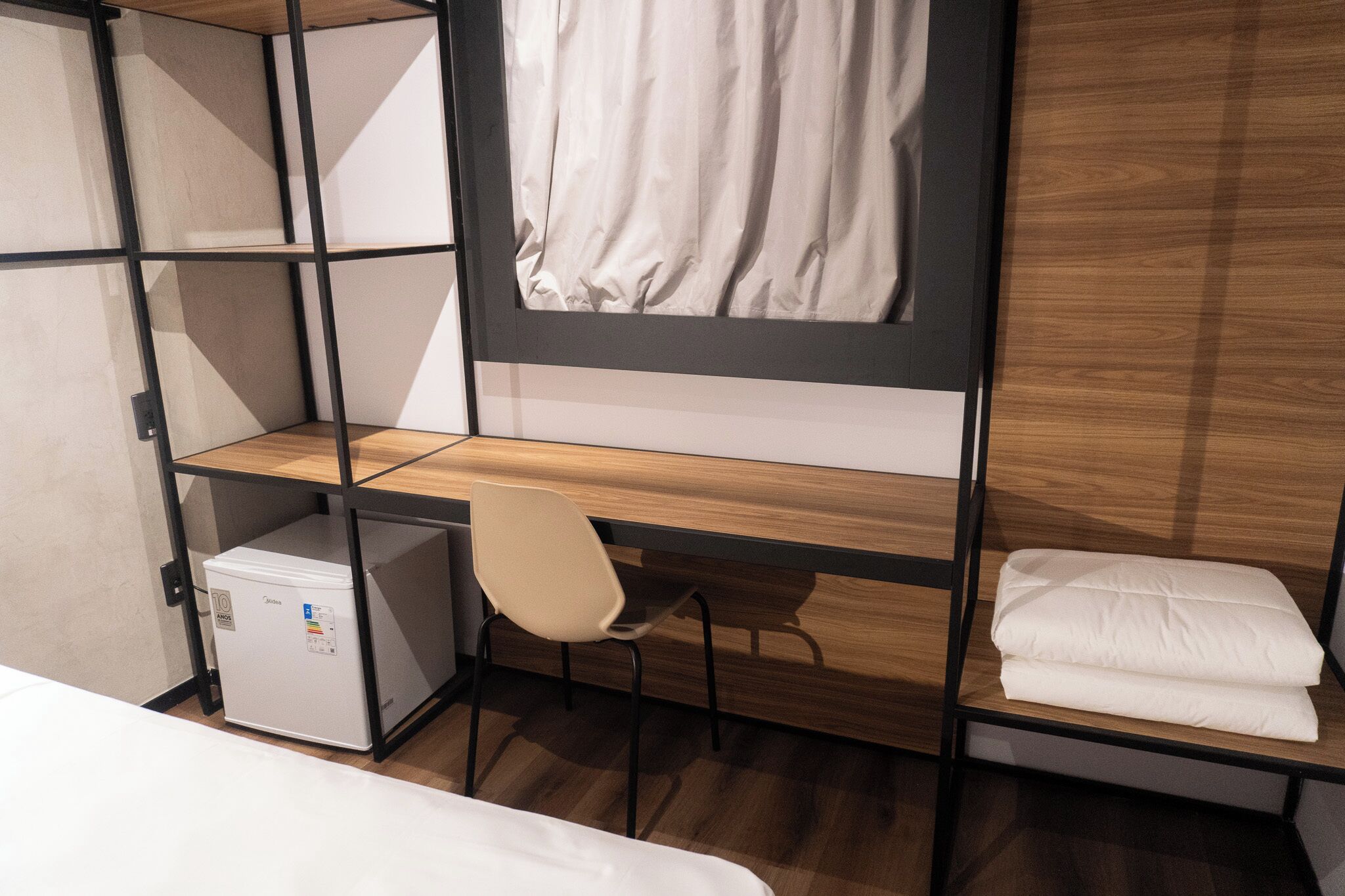 A compact room with a bunk bed, a small desk with a chair, a compact refrigerator, and a neatly folded white blanket.