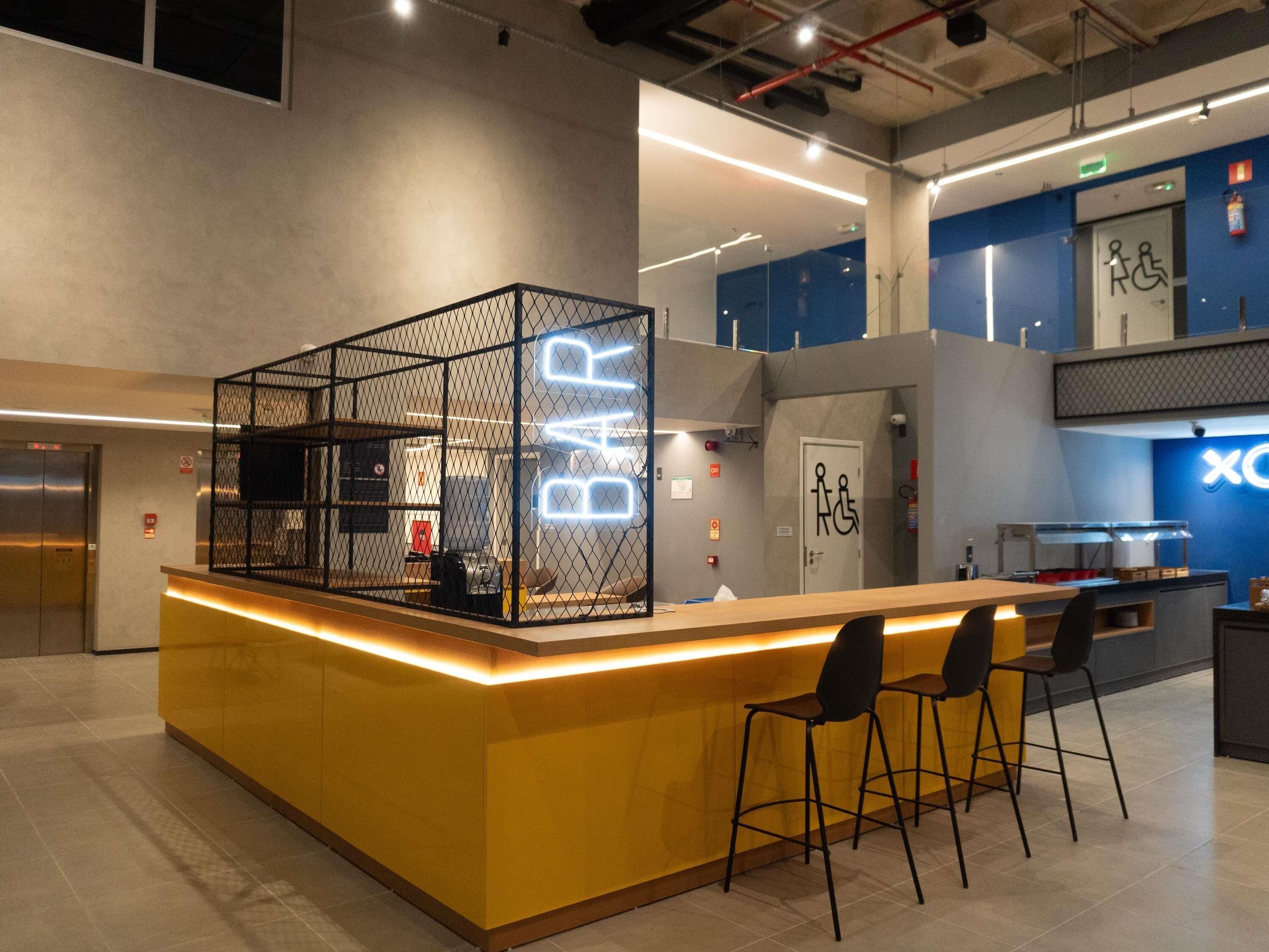Un área de bar moderna con mostrador amarillo, sillas negras y un letrero de neón.