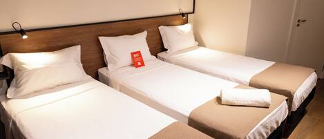 A hotel room with three single beds, each with white linens and brown accents. There is a wooden headboard, a wall-mounted light, and a door with an air conditioning unit.