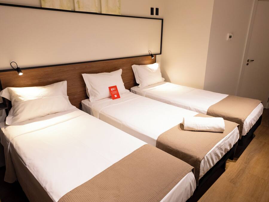 Ein Hotelzimmer mit drei Einzelbetten, jeweils mit weißer Bettwäsche und braunen Akzenten. Es gibt ein hölzernes Kopfteil, eine Wandleuchte und eine Tür mit Klimaanlage.