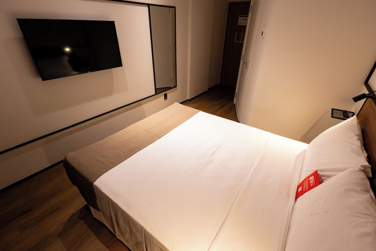 A hotel room with a bed, a flat-screen TV mounted on the wall, and a wooden floor.