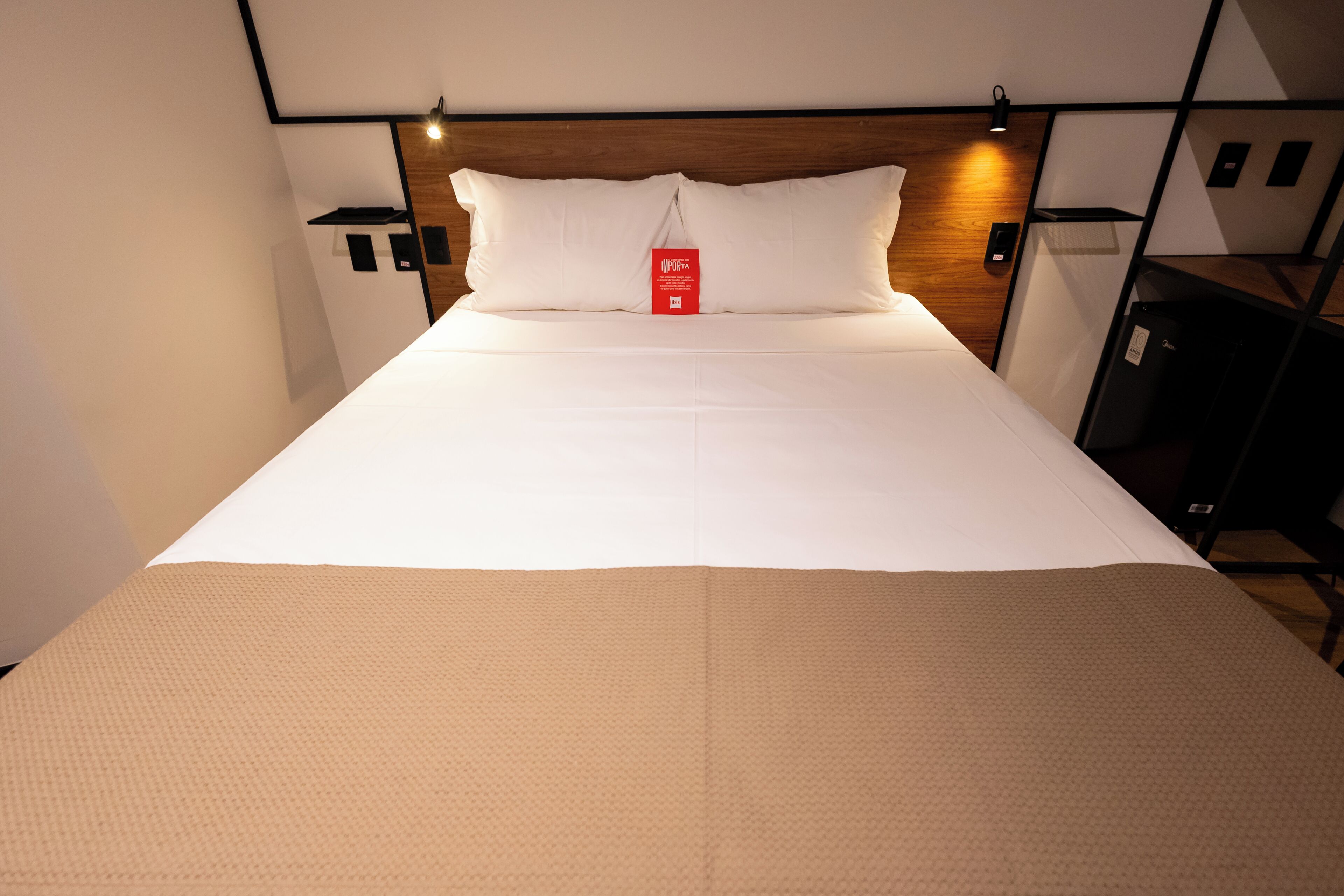 Una cama bien tendida con sábanas blancas y un plaid color beige, un cabecero de madera y una etiqueta roja en las almohadas.