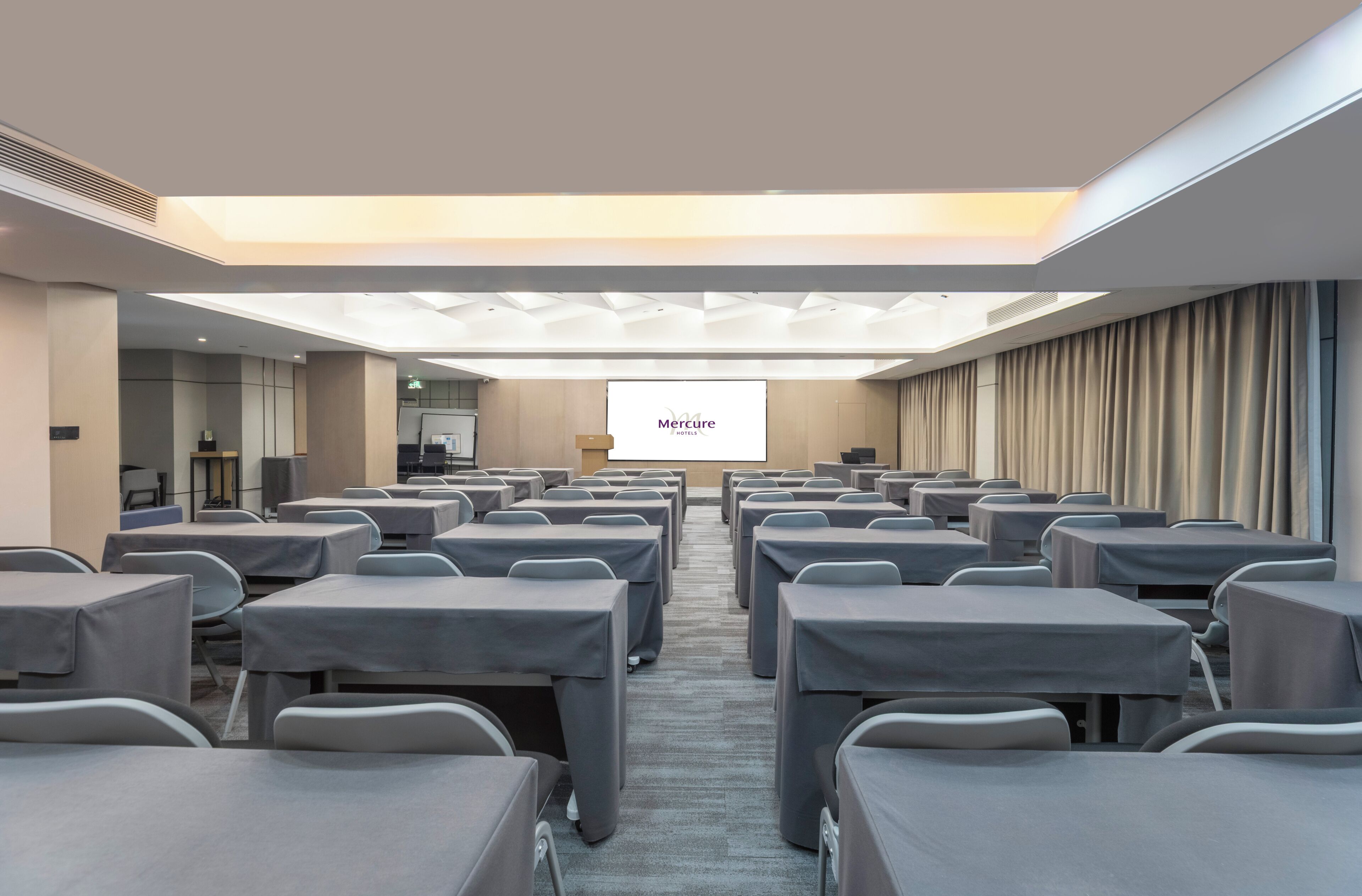 A conference room with rows of tables and chairs, a projection screen, and a visible brand name 'Mercure'.