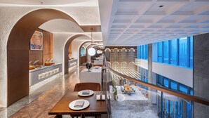 Un moderno vestíbulo de hotel con zona de comedor, mostrador de recepción y vistas a la ciudad a través de amplios ventanales.