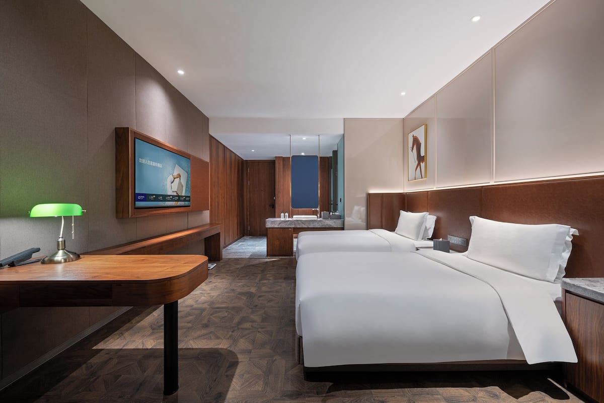 A hotel room with a large bed, a desk with a lamp, a TV mounted on the wall, and a bathroom visible through an open door.