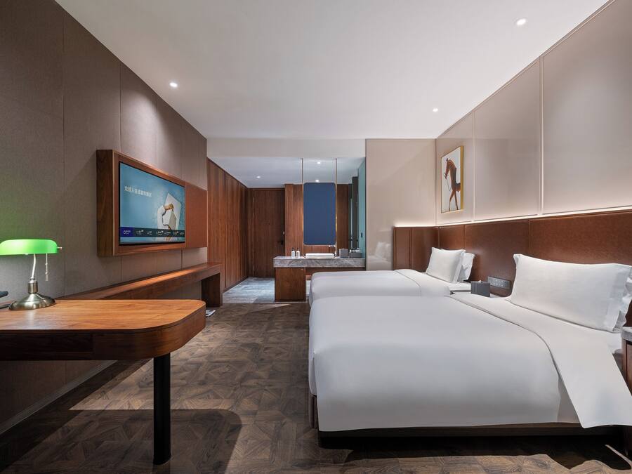 Una habitación de hotel con una cama grande, un escritorio con lámpara, un televisor montado en la pared y un baño visible a través de una puerta abierta.