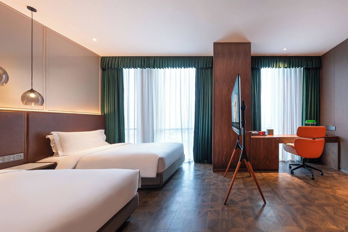 A hotel room with two beds, a desk with a chair, a large window with curtains, and a painting on the wall.