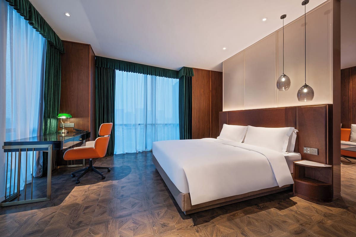 A hotel room with a large bed, a desk with a chair, and a view of the city through large windows.