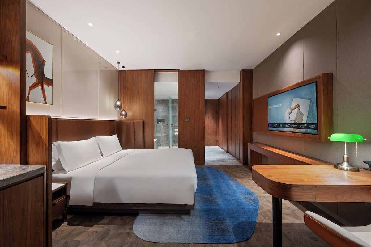 A modern hotel room with a large bed, a desk with a lamp, a TV, and a wooden wall panel.