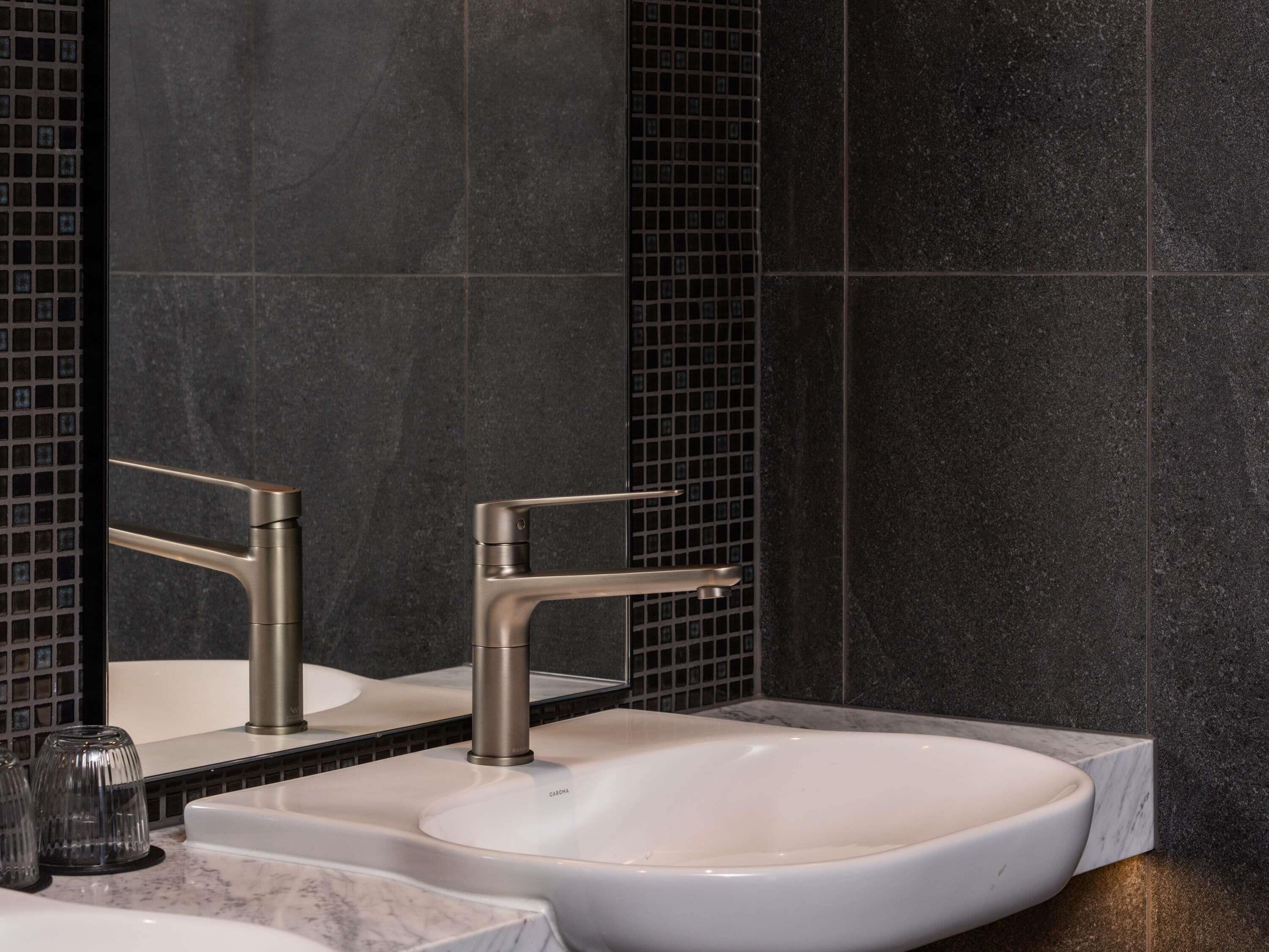 A bathroom with a white sink, a modern faucet, and a mirror with a black tiled backsplash.