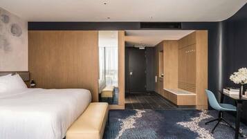 Una habitación de hotel moderna con una cama grande, una zona de estar, un escritorio y un baño visible a través de un separador de vidrio.