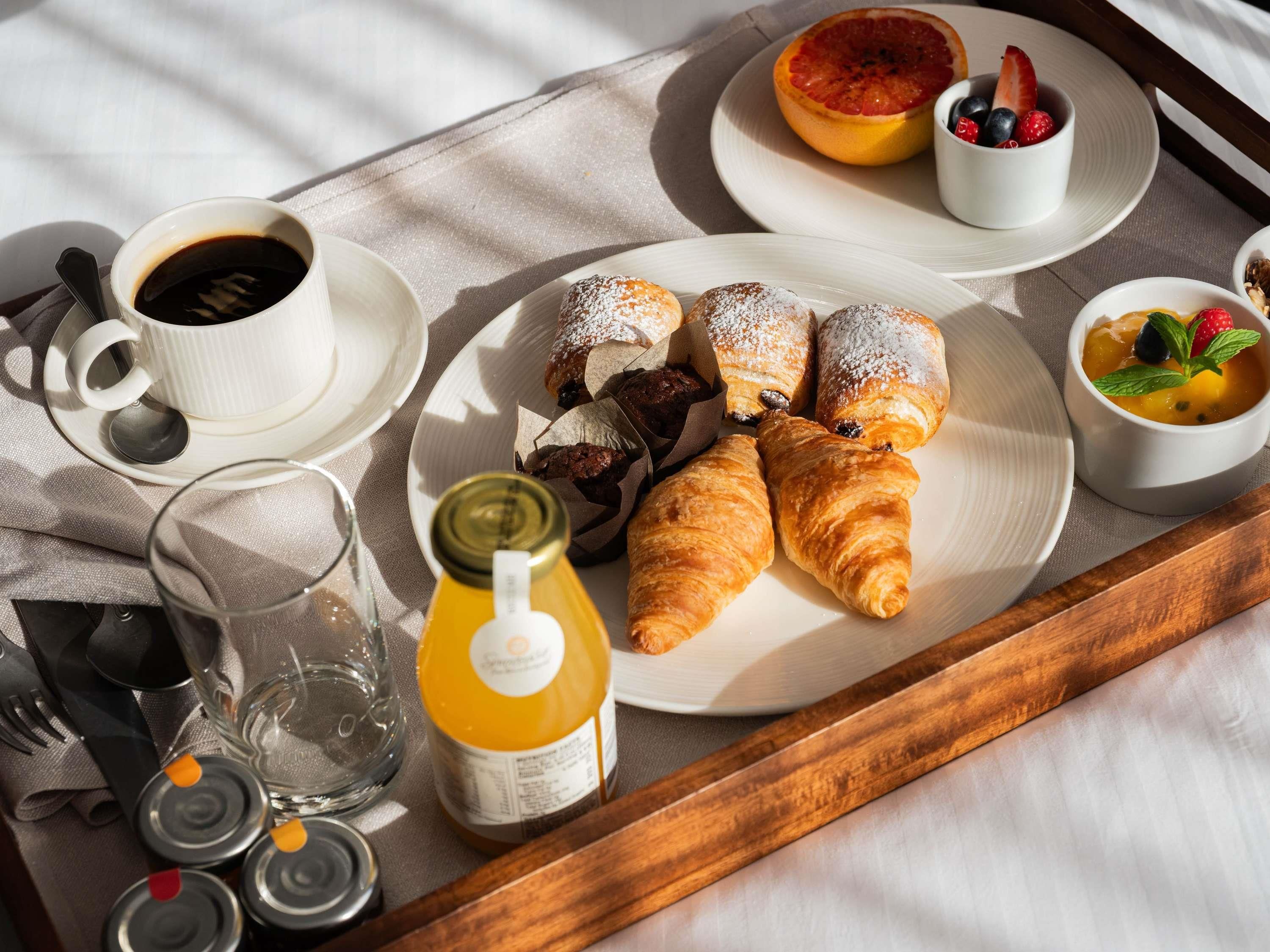 Bandeja de desayuno con cruasanes, una taza de café, un vaso de agua, una botella de jugo y un plato con fruta y pasteles.