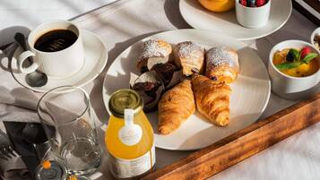Bandeja de desayuno con cruasanes, una taza de café, un vaso de agua, una botella de jugo y un plato con fruta y pasteles.