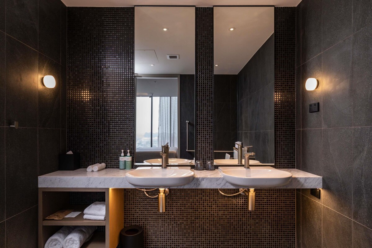 A modern bathroom with two sinks, a mirror, and a view of the city.