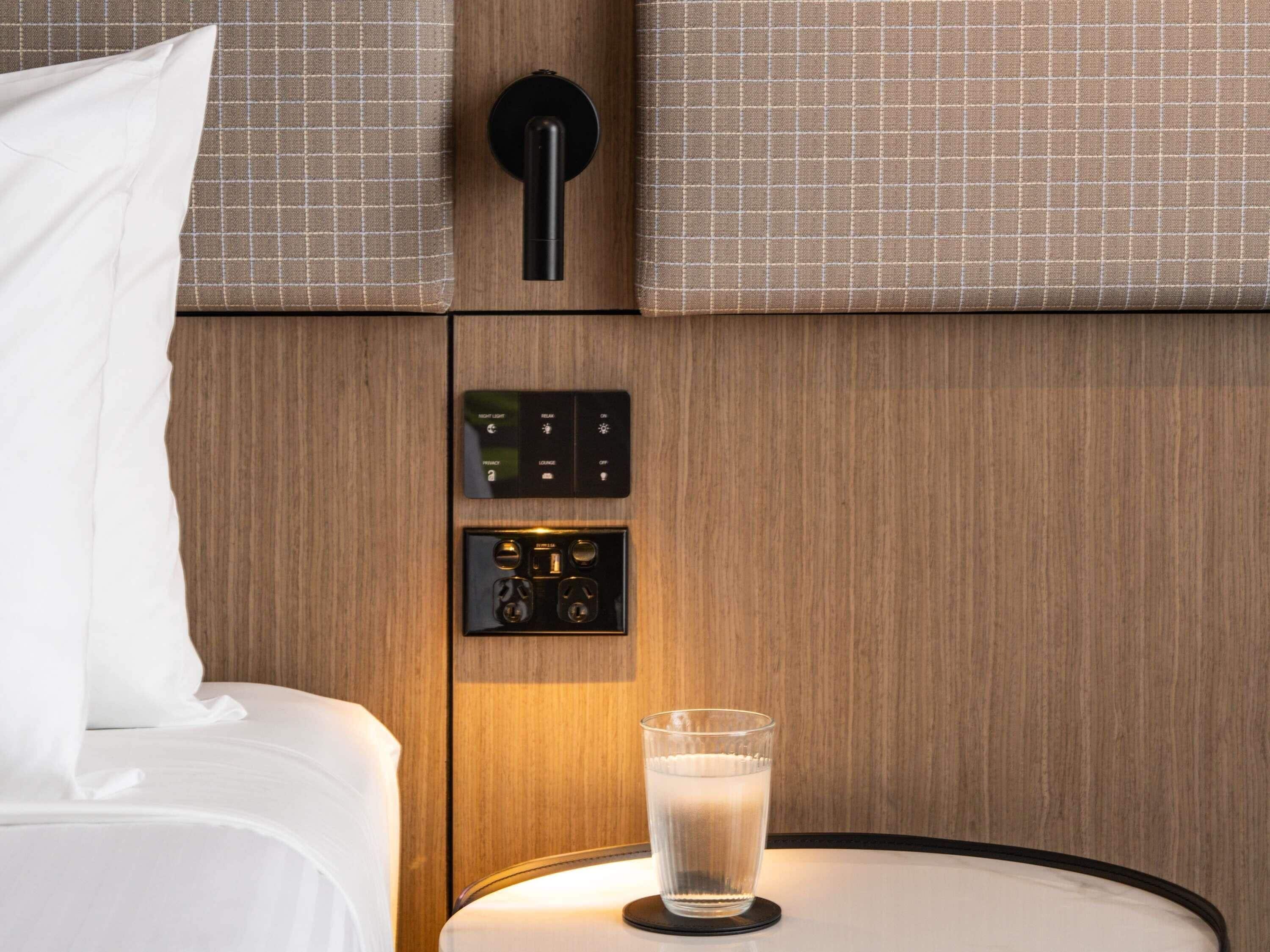 Een hotelkamer met een bed, een nachtkastje met een glas water, een bedieningspaneel aan de muur met knoppen, en een deur met een ronde hendel.