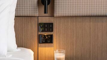 Een hotelkamer met een bed, een nachtkastje met een glas water, een bedieningspaneel aan de muur met knoppen, en een deur met een ronde hendel.