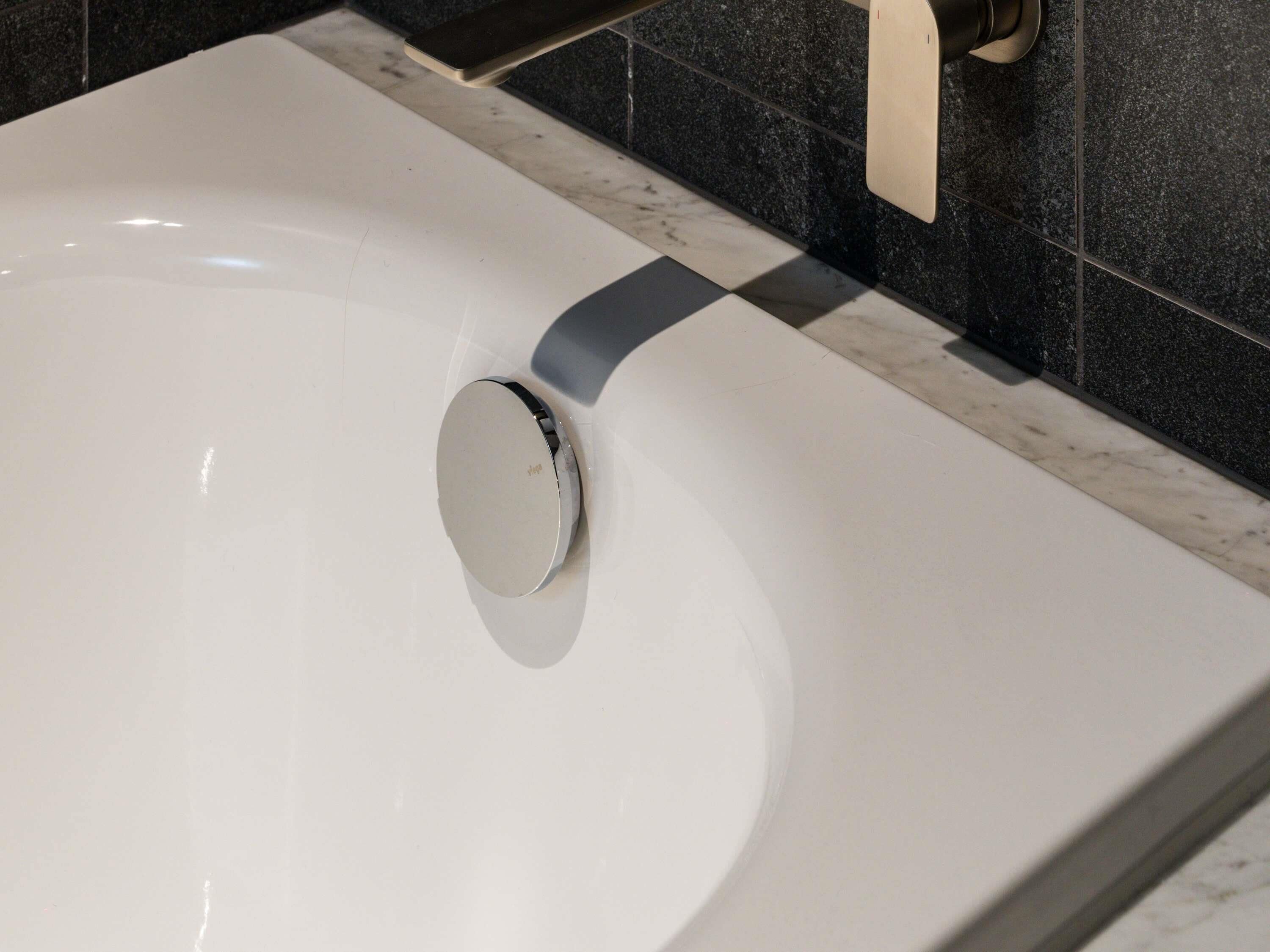 Un évier blanc doté d’un robinet moderne, un plan de travail en marbre et des murs recouverts de carreaux foncés.