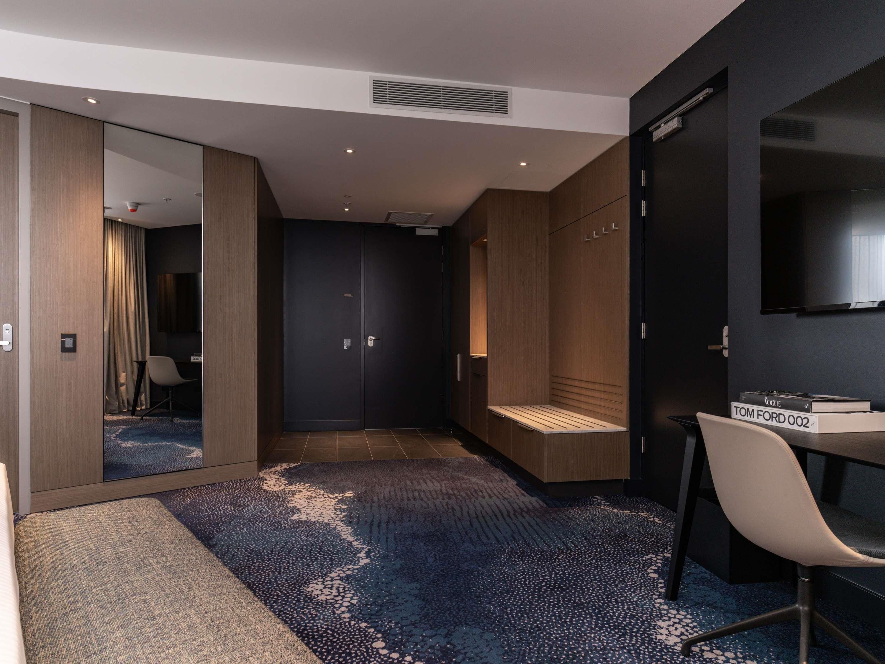 Quarto de hotel moderno com um espelho grande, televisão, secretária com cadeira e guarda-roupa.