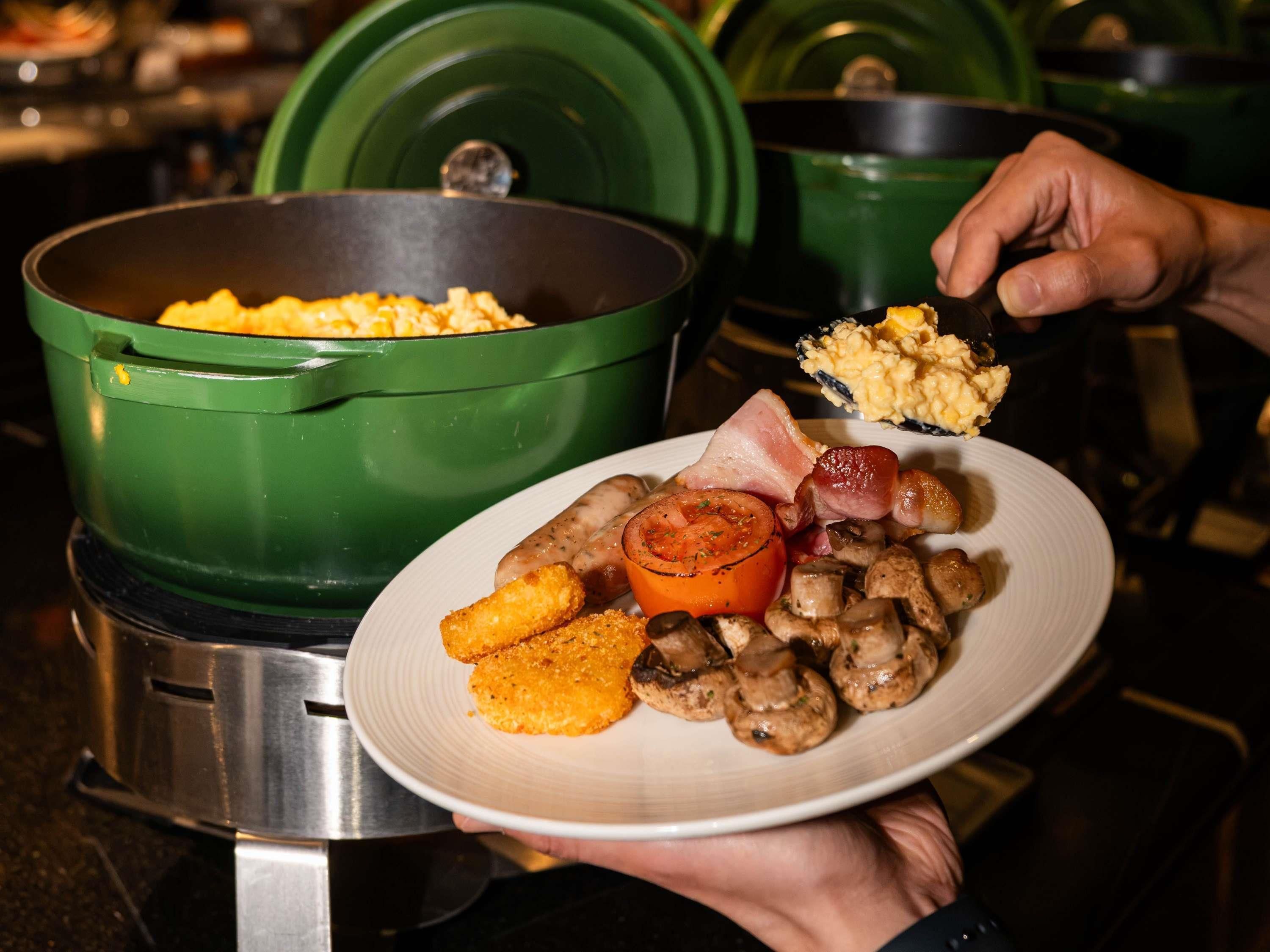 Una persona sostiene un plato con comida, incluyendo salchichas, champiñones y un tomate. Al fondo, hay una olla verde sobre una estufa.
