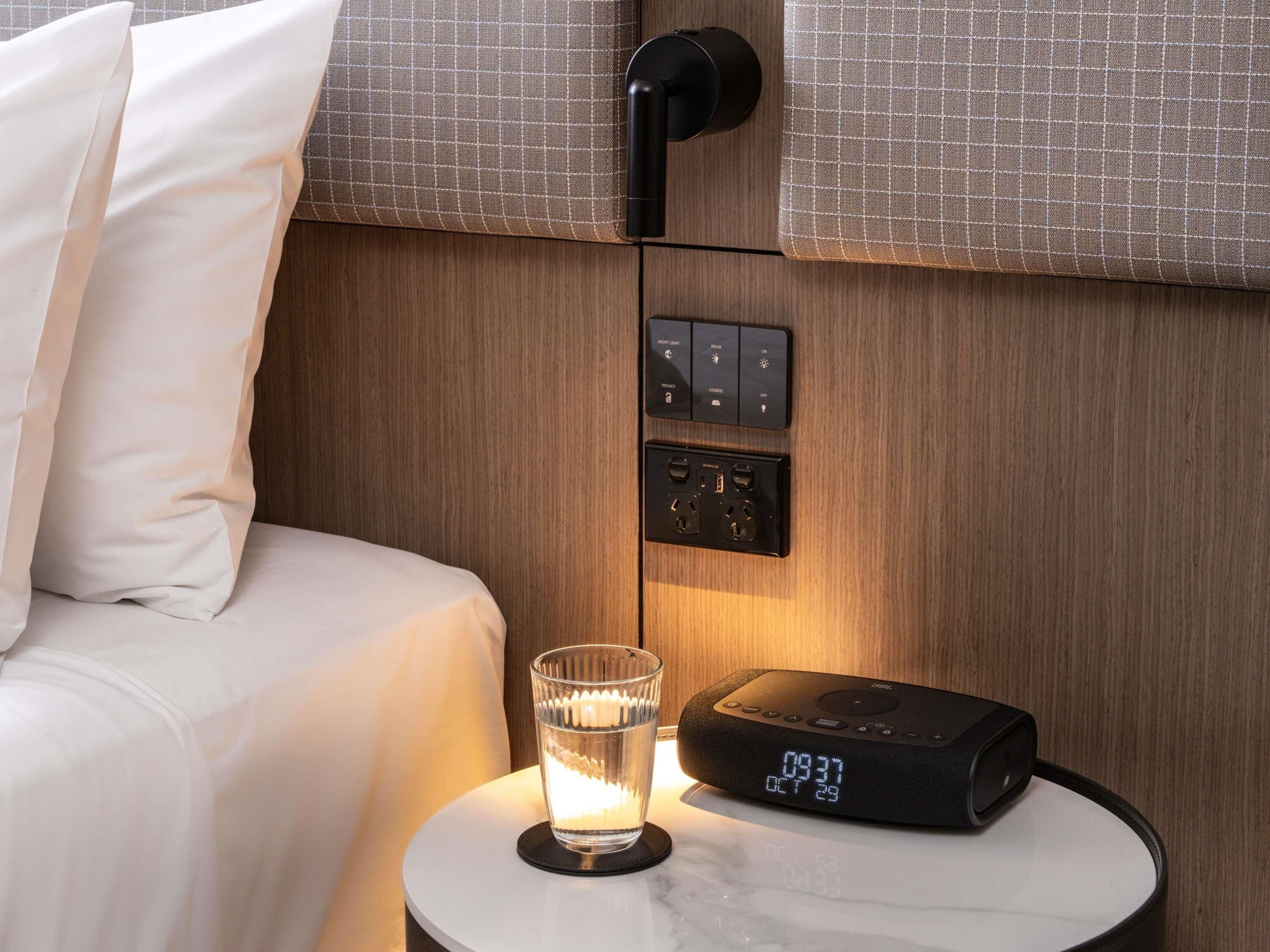 A hotel room with a bed, a bedside table with a clock, a glass of water, and a wall-mounted control panel.