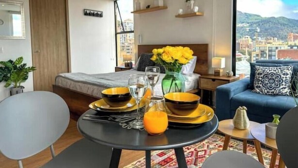 A modern hotel room with a bed, a dining table set with yellow plates and glasses, a vase with yellow flowers, and a view of the city through large windows. - Beautiful Loft in Bogota-Charming View-Gym (Bogotá)