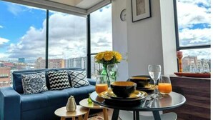 A modern living room with a blue sofa, a round dining table with orange juice and bowls, and a large window with a city view. - Beautiful Loft in Bogota-Charming View-Gym (Bogotá)