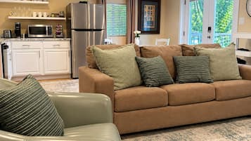 A living room with a beige sofa, a green armchair, and a coffee table. There is a kitchen area with white cabinets, a refrigerator, and a microwave.