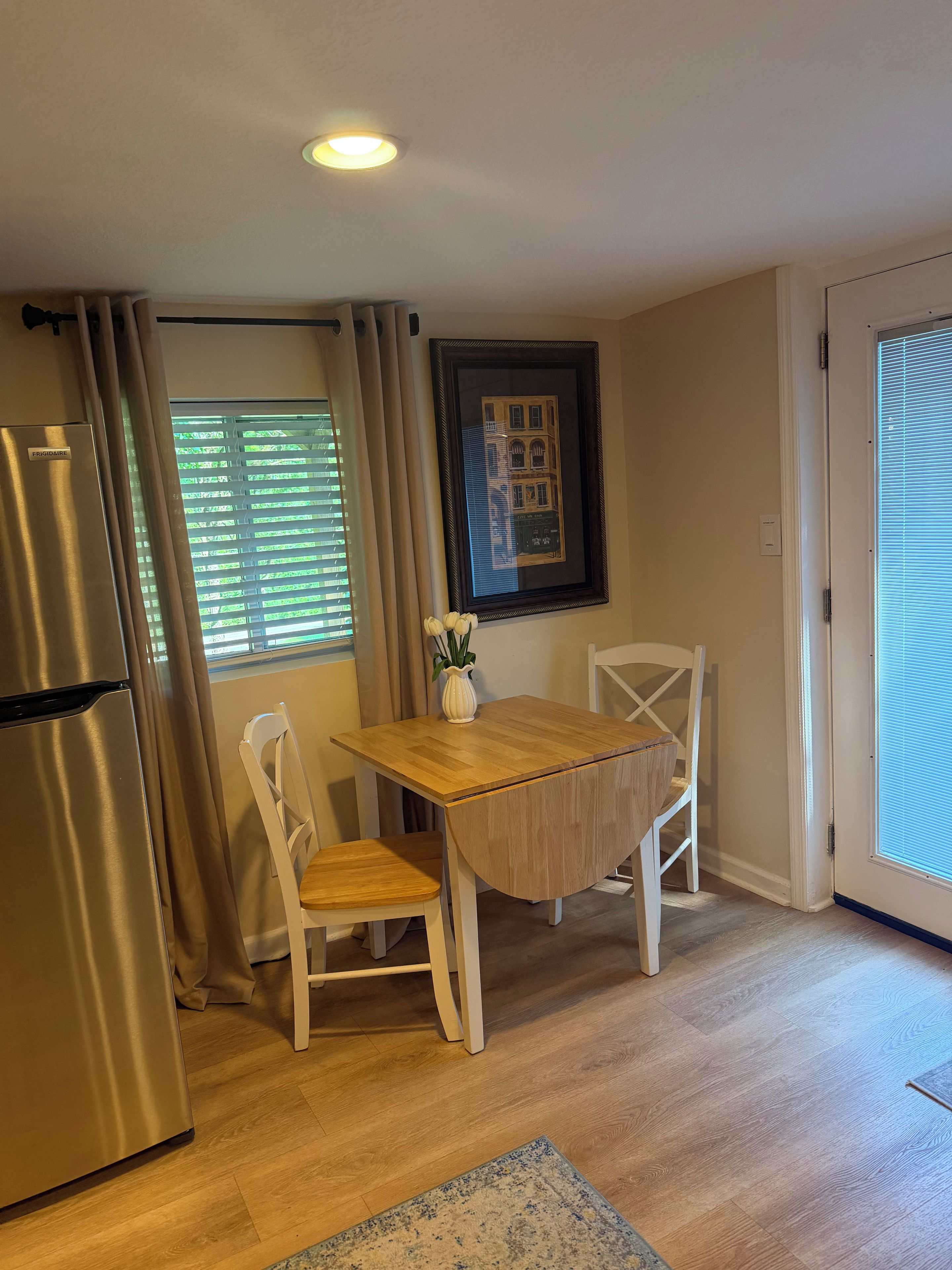 Eine kleine Küche mit Kühlschrank, einem Esstisch mit Stühlen, einem gerahmten Bild an der Wand und einem Fenster mit Jalousien.