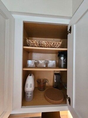 A kitchen cabinet with a woven basket, measuring cups, a coffee maker, a grater, and a cutting board.