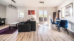 A modern living room with a black sofa, a coffee table, and a dining area with blue chairs. There is a large window and a door leading to a balcony.
