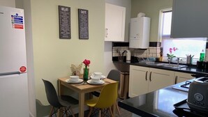 A compact kitchen with a dining area, featuring a refrigerator, a coffee machine, and a small dining table with chairs. - Buccleuch Whole Apartment (Kettering)