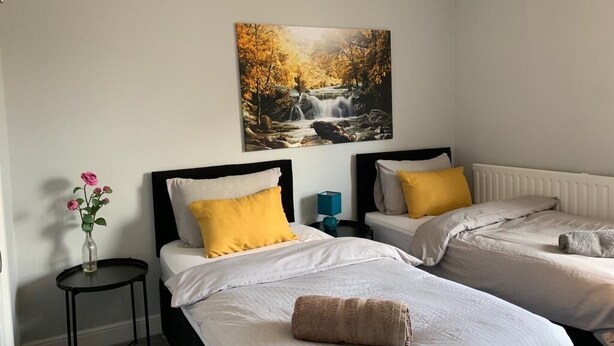 A hotel room with two beds, a painting of a waterfall, a vase with flowers, and a small table. - Buccleuch Whole Apartment (Kettering)