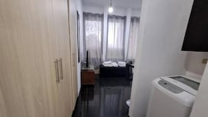 A hotel room with a wooden wardrobe, a small table with a lamp, a bed with white linens, a washing machine, and a window with sheer curtains. - Suite in San Miguel Steps from Monagas Plaza (Monagas)