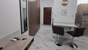 Een moderne kamer met een houten bureau, twee zwarte stoelen, een spiegel, een ronde wandklok en een kitchenette met houten kastjes en een koelkast.