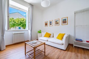 A modern living room with a white sofa, a small table, and a radiator. There are three framed pictures on the wall and a window with curtains.