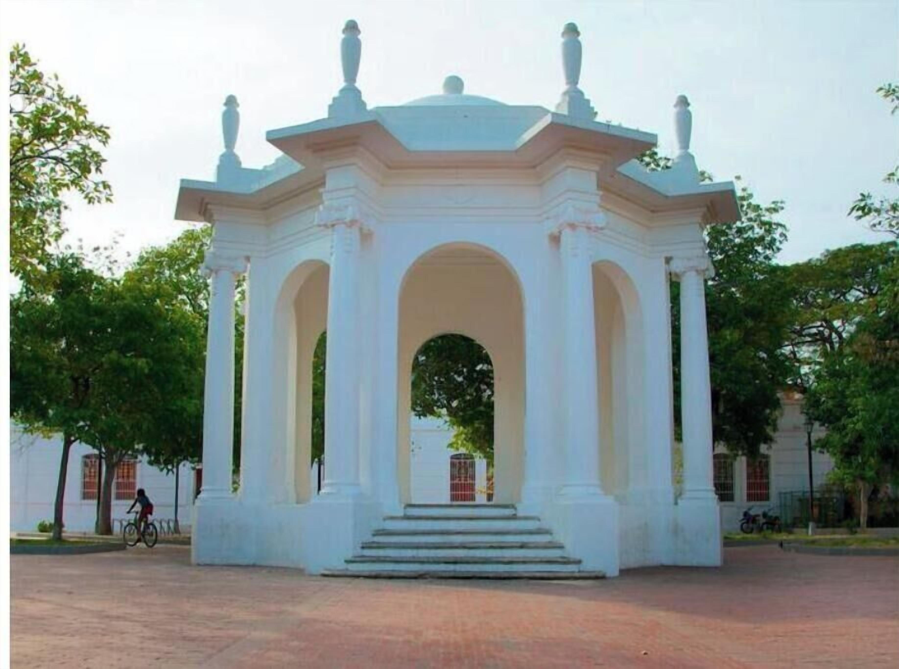 A white gazebo with a central archway and steps leading up to it, surrounded by trees.
