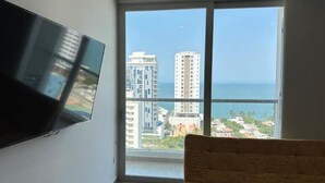 A room with a large window, a flat-screen TV mounted on the wall, and a view of high-rise buildings and the ocean.
