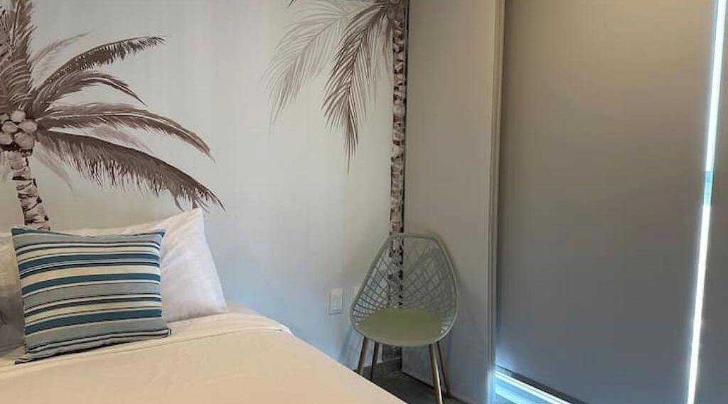 Une chambre à coucher avec un lit, un palmier décoratif, un coussin à rayures et une chaise moderne.