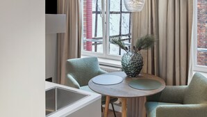 Un'area pranzo moderna con un tavolo rotondo in legno, due sedie verdi, un vaso decorativo con rami di pino e vista su un paesaggio innevato attraverso una finestra con tende beige.