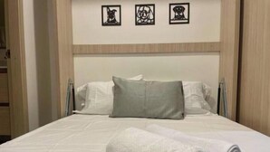 Un lit bien fait, avec du linge de lit blanc et un oreiller gris, une serviette blanche enroulée, et trois tableaux encadrés accrochés au mur au-dessus.