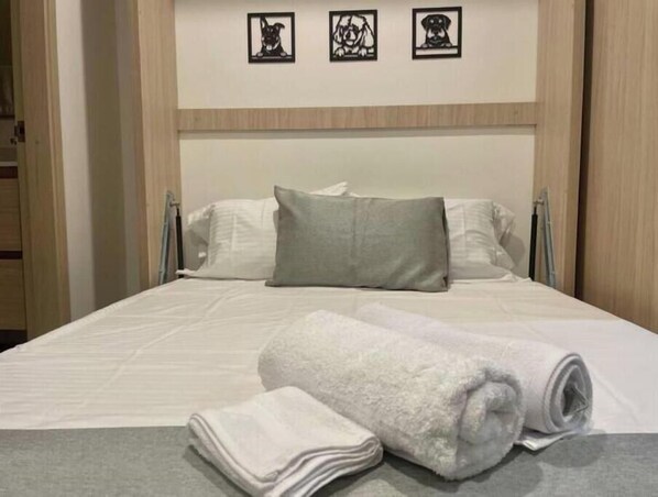 A neatly made bed with white linens and a grey pillow, a rolled white towel, and three framed pictures on the wall above.