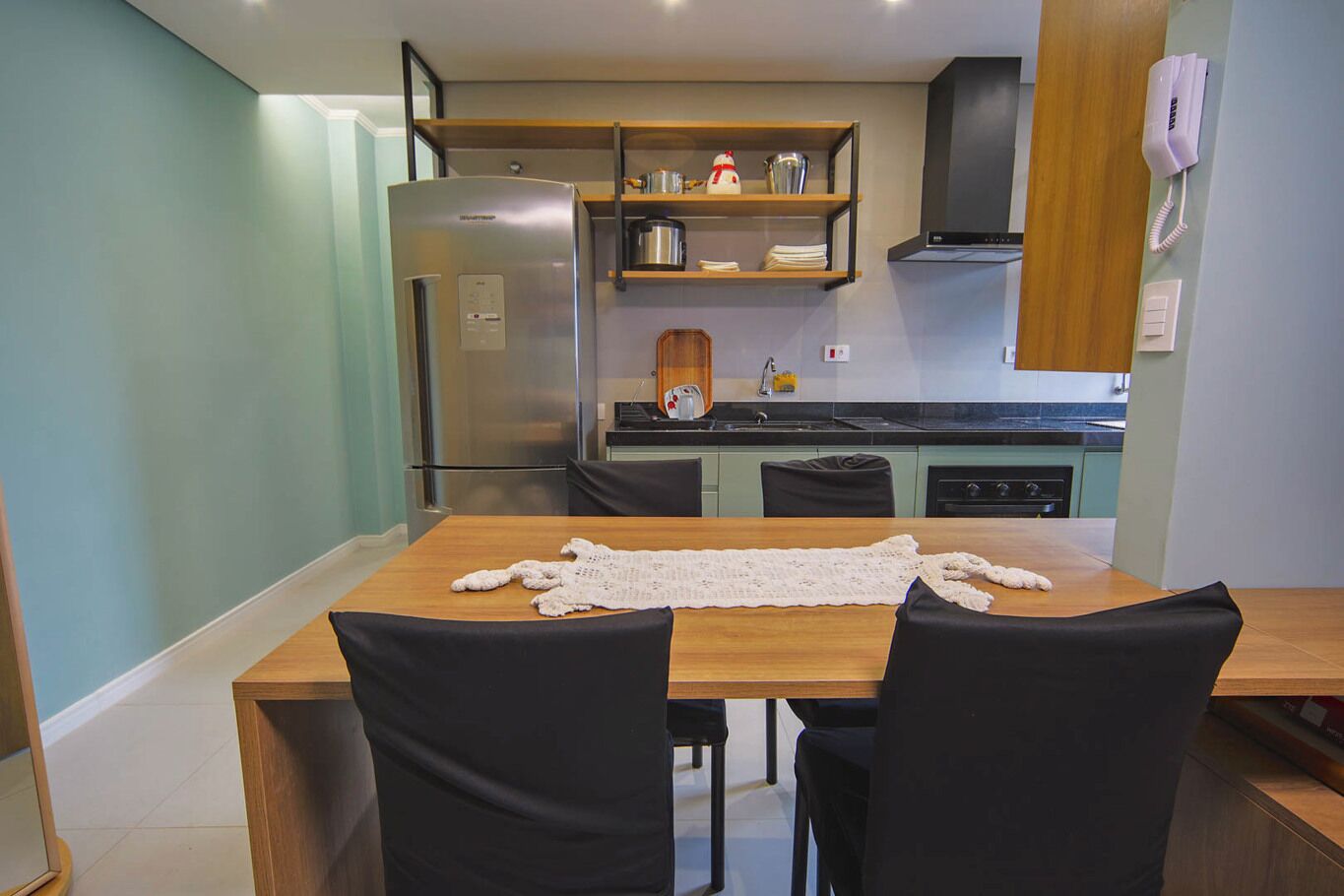 A modern kitchen with a stainless steel refrigerator, a wooden table with a crocheted placemat, and black chairs.