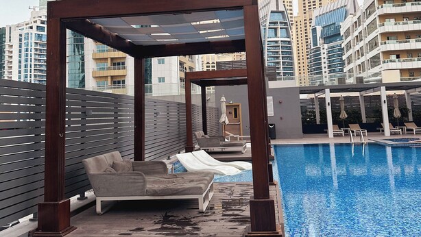 Ein Poolbereich auf dem Dach mit einer Holzpergola, Liegestühlen und Blick auf Gebäude der Stadt im Hintergrund.
