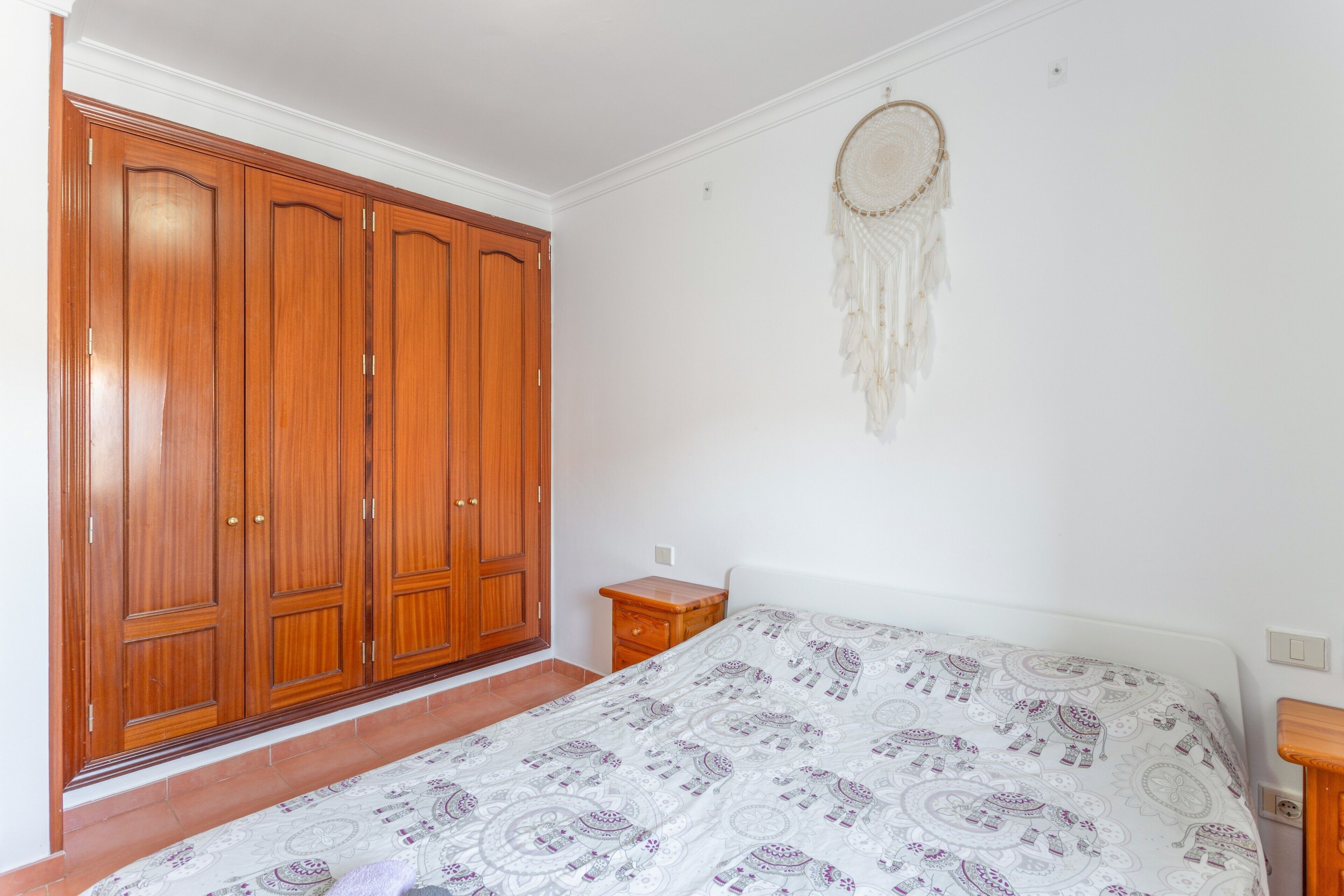 Une chambre avec une grande armoire en bois, un lit avec une literie à motifs et un attrape-rêves accroché au mur.