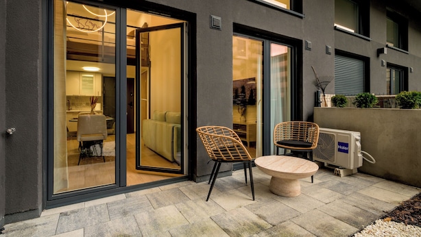 A modern building exterior with a glass door entrance, a dining area with wicker chairs and a round table, and an air conditioning unit on the right. - RentPlanet - Sienkiewicza Residence (Wrocław)