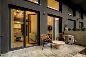 A modern building exterior with a glass door entrance, a dining area with wicker chairs and a round table, and an air conditioning unit on the right. - RentPlanet - Sienkiewicza Residence (Wrocław)