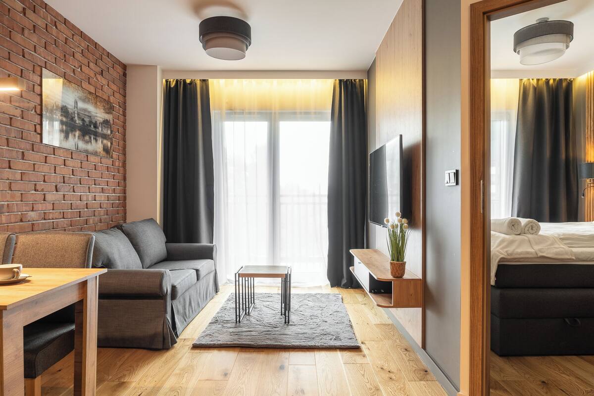 A modern hotel room with a brick accent wall, a sofa, a small table, a TV, and a bed.