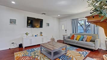 Una sala de estar con un televisor de pantalla plana montado en la pared, un sofá gris con cojines coloridos, una mesa de centro de madera y una alfombra estampada.
