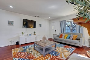 A living room with a flat-screen TV mounted on the wall, a grey sofa with colorful cushions, a wooden coffee table, and a patterned rug.
